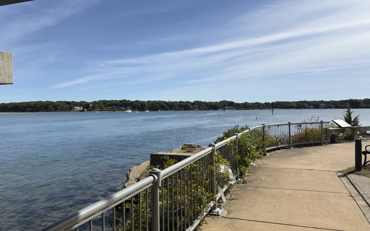 Curving waterfront walkway with a railing and a bench beside Lagoon Pond, wooded shore and moored boats across the water under a blue sky