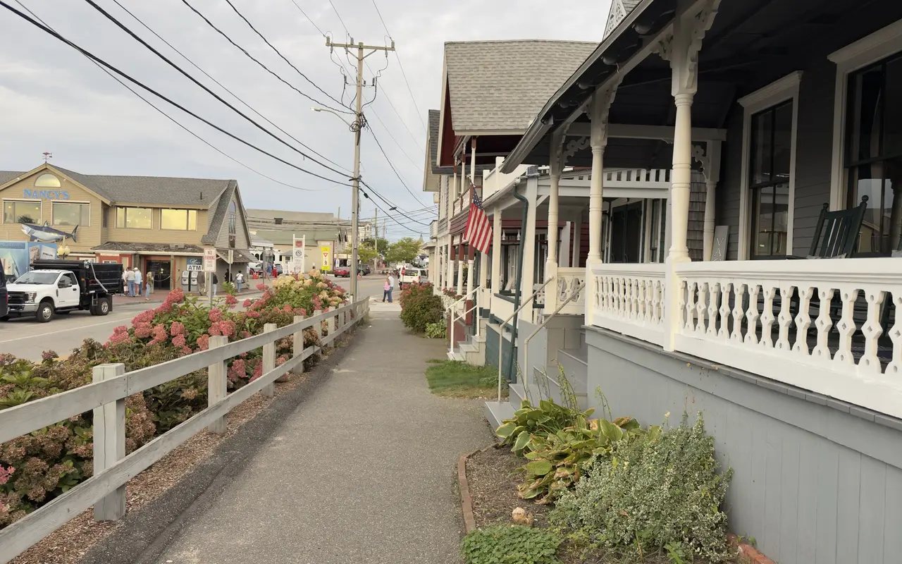 A sidewalk along Lake Avenue in Oak Bluffs: gingerbread cottage porches with white cutout railings, rocking chairs and a Vacancy sign on the right, pink hydrangeas along a wooden fence on the left, and Nancy's restaurant with its shark sign across the street