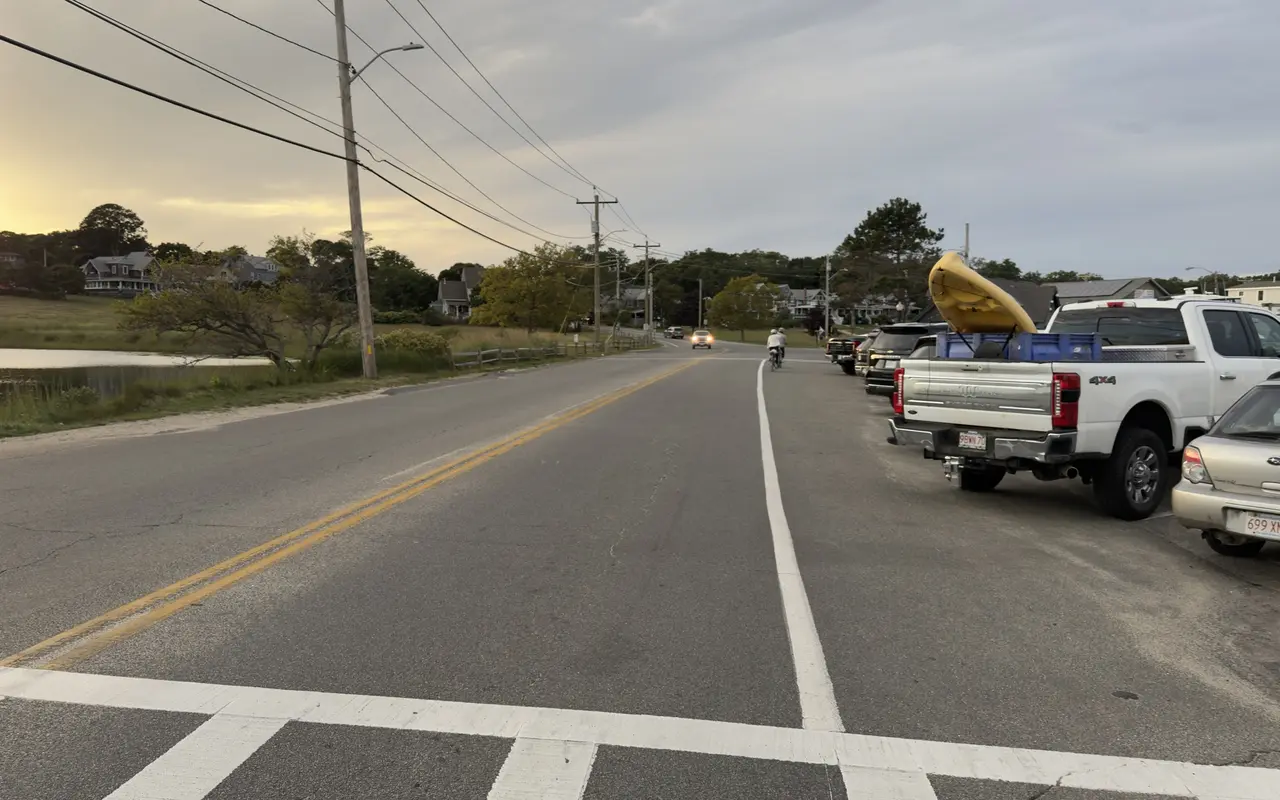 Lake Avenue in Oak Bluffs at sunset: Sunset Lake and marsh grass on the left, a double yellow line, a marked bike lane with two cyclists, cars parked along the right with a yellow kayak in a pickup bed, a crosswalk in the foreground