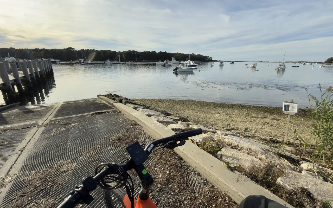 The handlebars of an orange e-bike at the top of the Lake Street boat ramp, Lake Tashmoo and its moored boats beyond