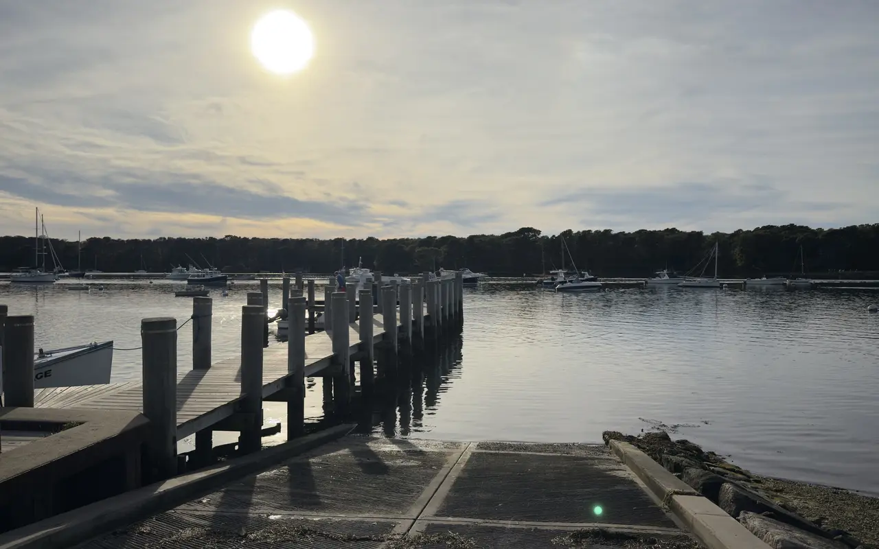 A wooden pier on pilings and a concrete boat ramp running into Lake Tashmoo, the sun low over moored boats and a wooded shore