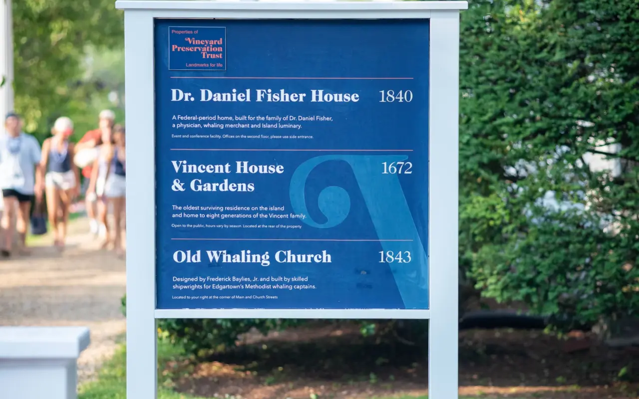 A blue Vineyard Preservation Trust sign in Edgartown listing the Dr. Daniel Fisher House, Vincent House and Gardens, and the Old Whaling Church