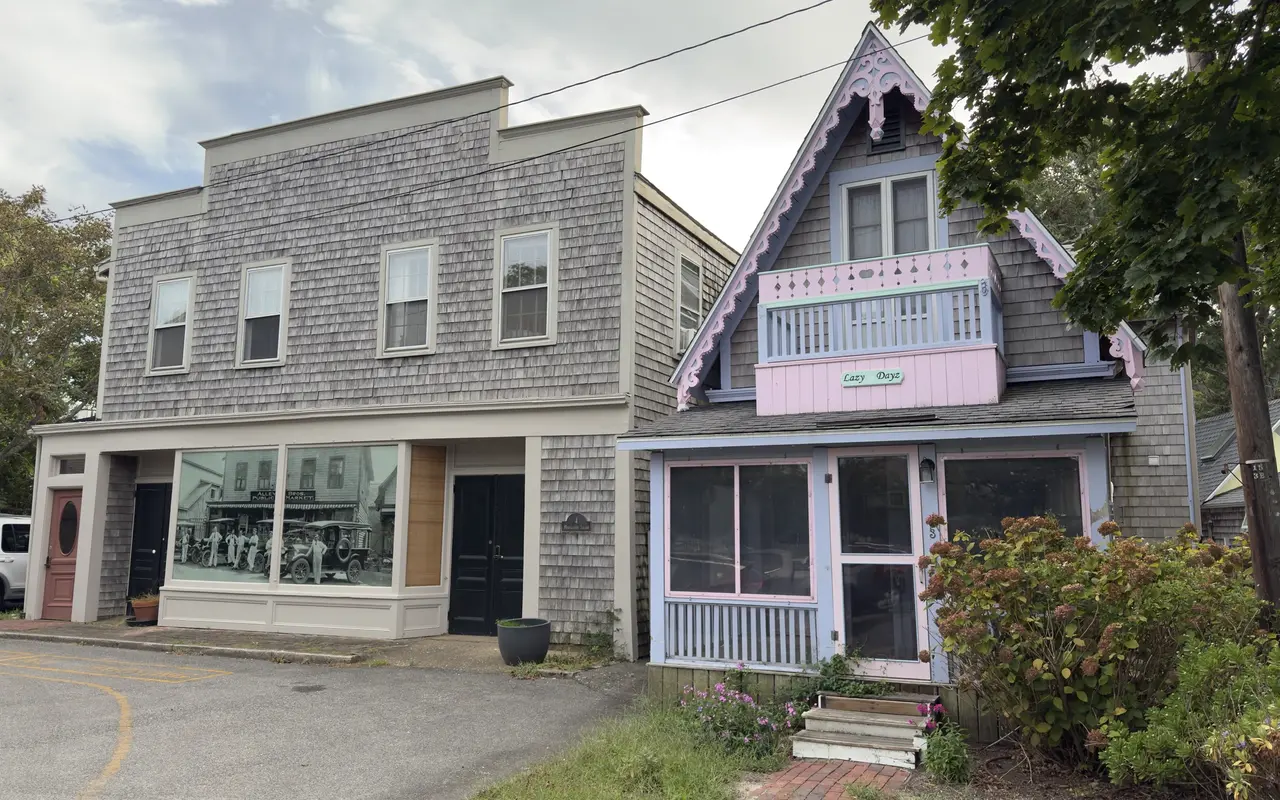 A small grey-shingled cottage called Lazy Dayz with pink and lavender trim and a screened porch, next to a grey shingled two-storey building whose shop window holds a large old black and white photograph of men and cars outside a public market