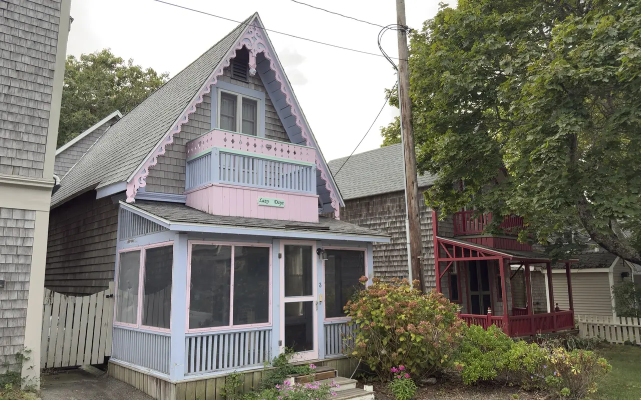 A grey-shingled gingerbread cottage called Lazy Dayz on Montgomery Square in Oak Bluffs with pink scrollwork, a lavender and mint balcony with heart cutouts, a screened porch, hydrangeas by the steps and a red-trimmed cottage next door