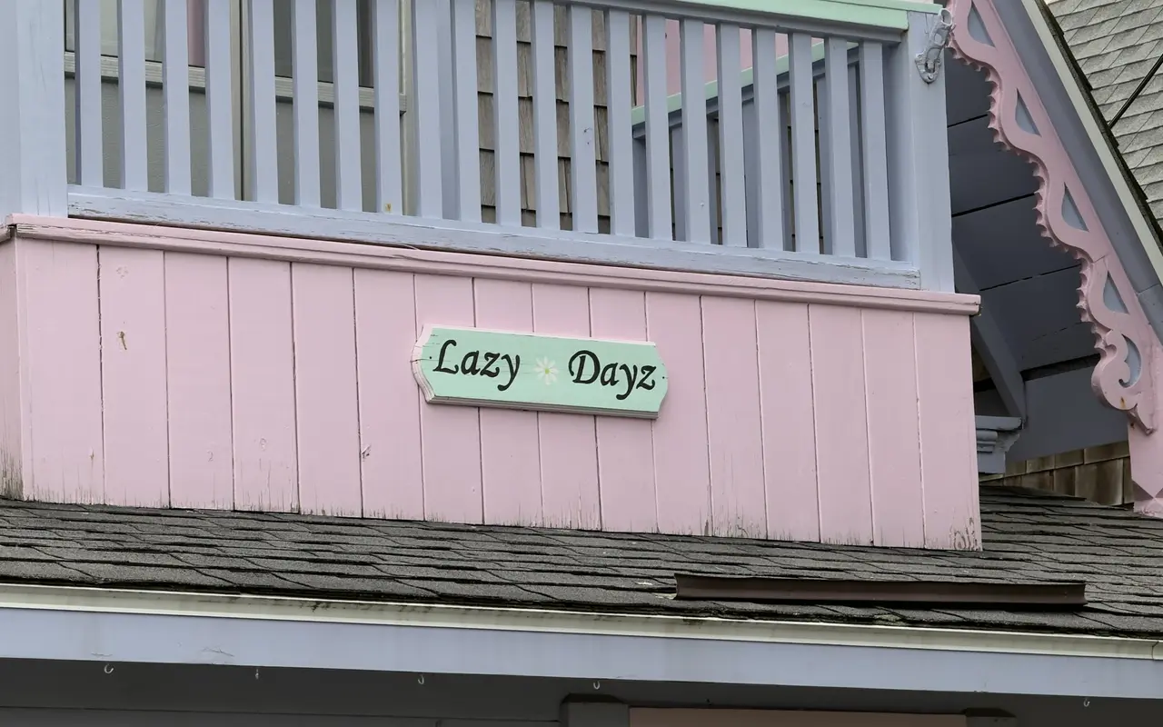 A small green name board reading Lazy Dayz with a daisy, on the pink skirt of a balcony with lavender railings and pink scrollwork on a gingerbread cottage in Oak Bluffs