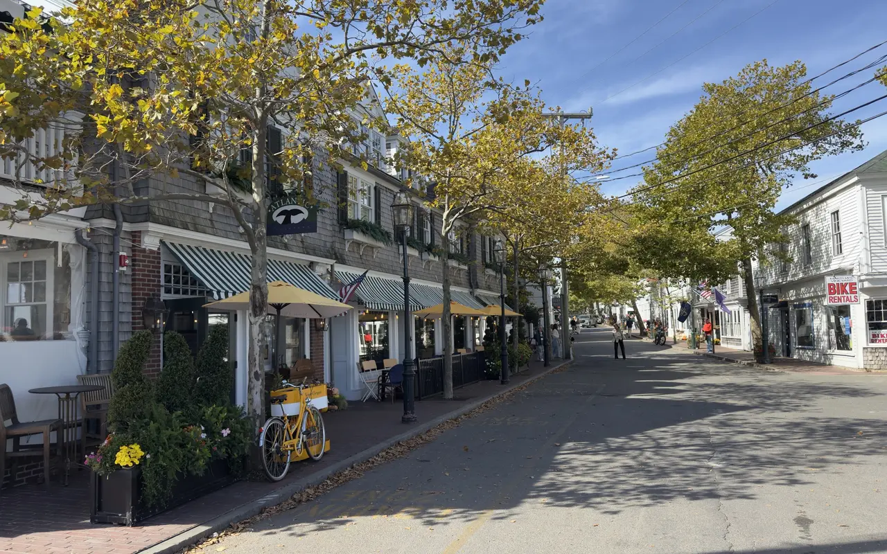 Lower Main Street in Edgartown with The Atlantic restaurant’s striped awnings and yellow umbrellas, a yellow bicycle by a planter, a bike rental shop opposite and plane trees in fall color