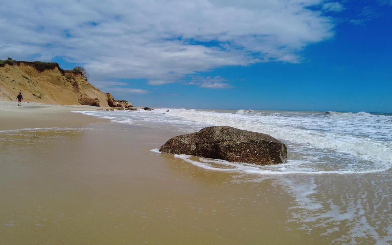 A large boulder in the shallow surf on a wide sandy beach, clay bluffs and two small walkers in the distance under a blue sky