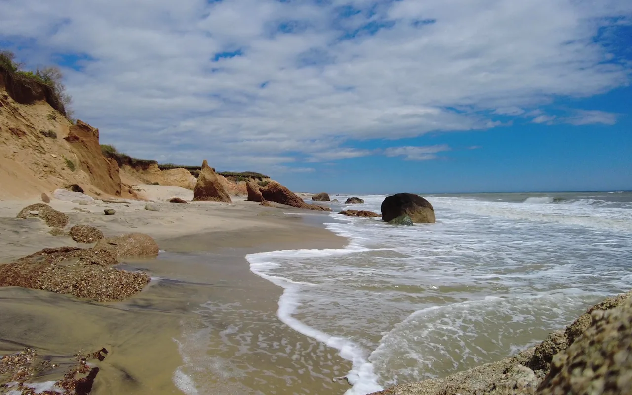 Eroded sandy and red clay bluffs above a beach scattered with boulders, waves washing in on the right