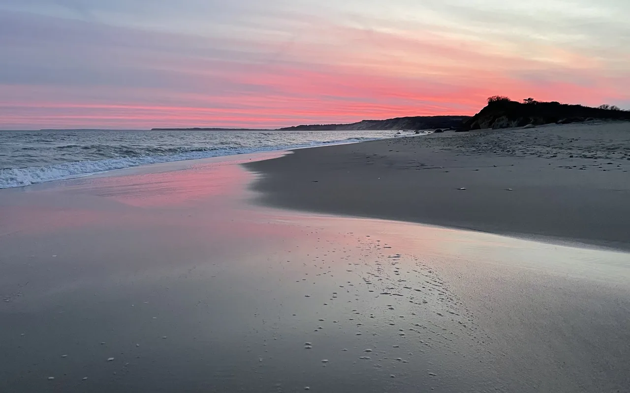 A wide empty beach at dusk with pink streaks of cloud reflected in the wet sand, small waves on the left and a dark bluff on the right