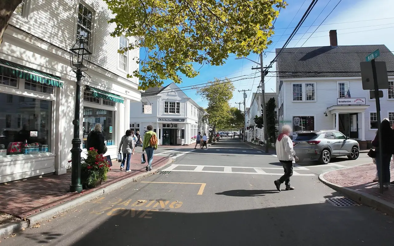 The corner of Main Street and Water Street in Edgartown with white clapboard shops, brick sidewalks, a crosswalk and people walking, street signs for Main St and N Water St