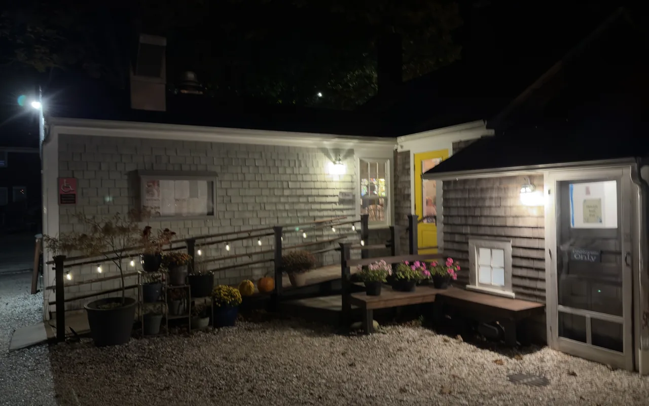 Grey-shingled Maker Café in Vineyard Haven at night with a yellow door, string lights along the railing and potted mums and pumpkins