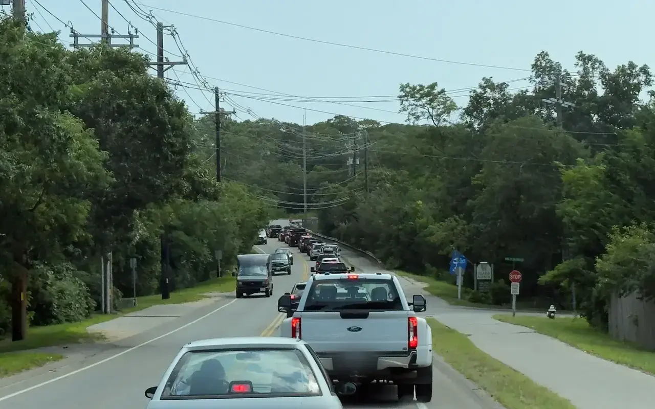 A line of cars and a white pickup backed up on a two-lane road through the trees on a summer day