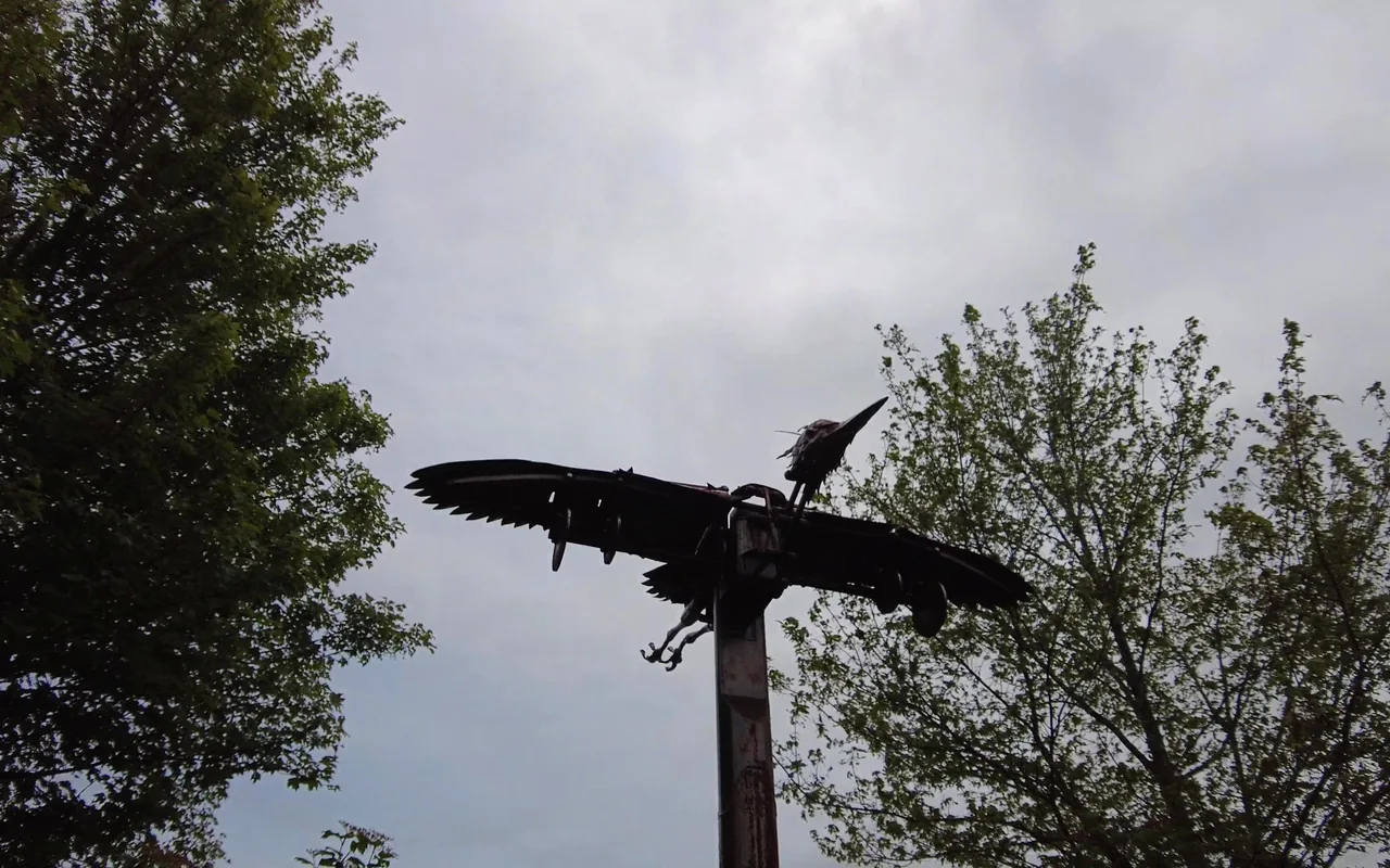 Dark metal sculpture of a bird with outstretched wings on top of a tall post, against a grey sky between trees