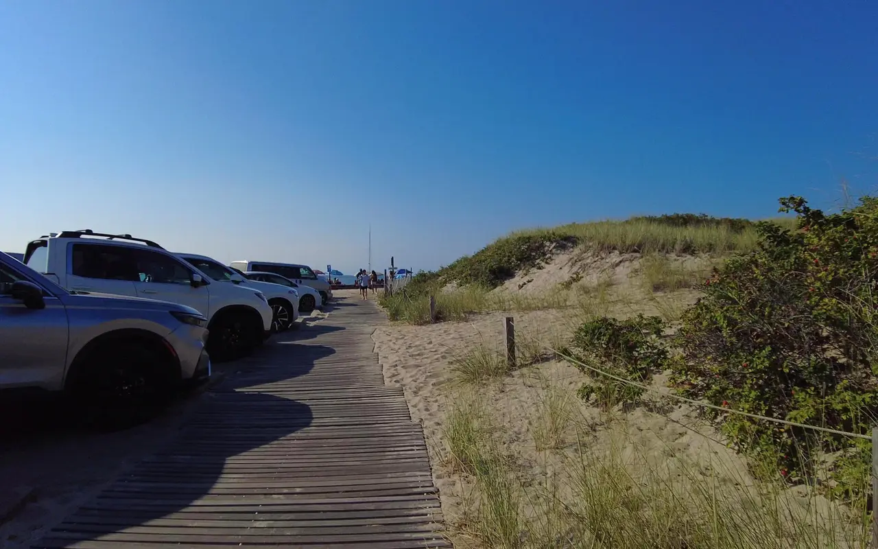 Weathered wooden boardwalk running between parked cars and a grassy dune toward Menemsha Beach on a sunny day