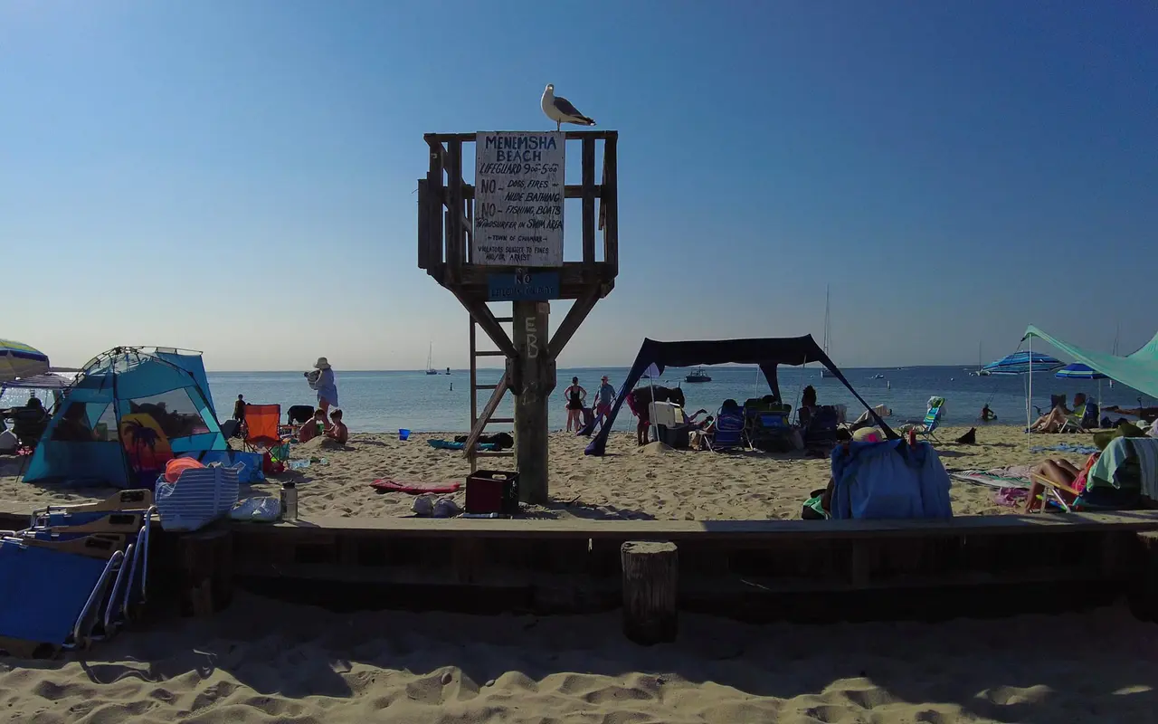 Wooden lifeguard stand with a gull perched on top and a hand-painted Menemsha Beach rules sign, beach tents, umbrellas and swimmers behind