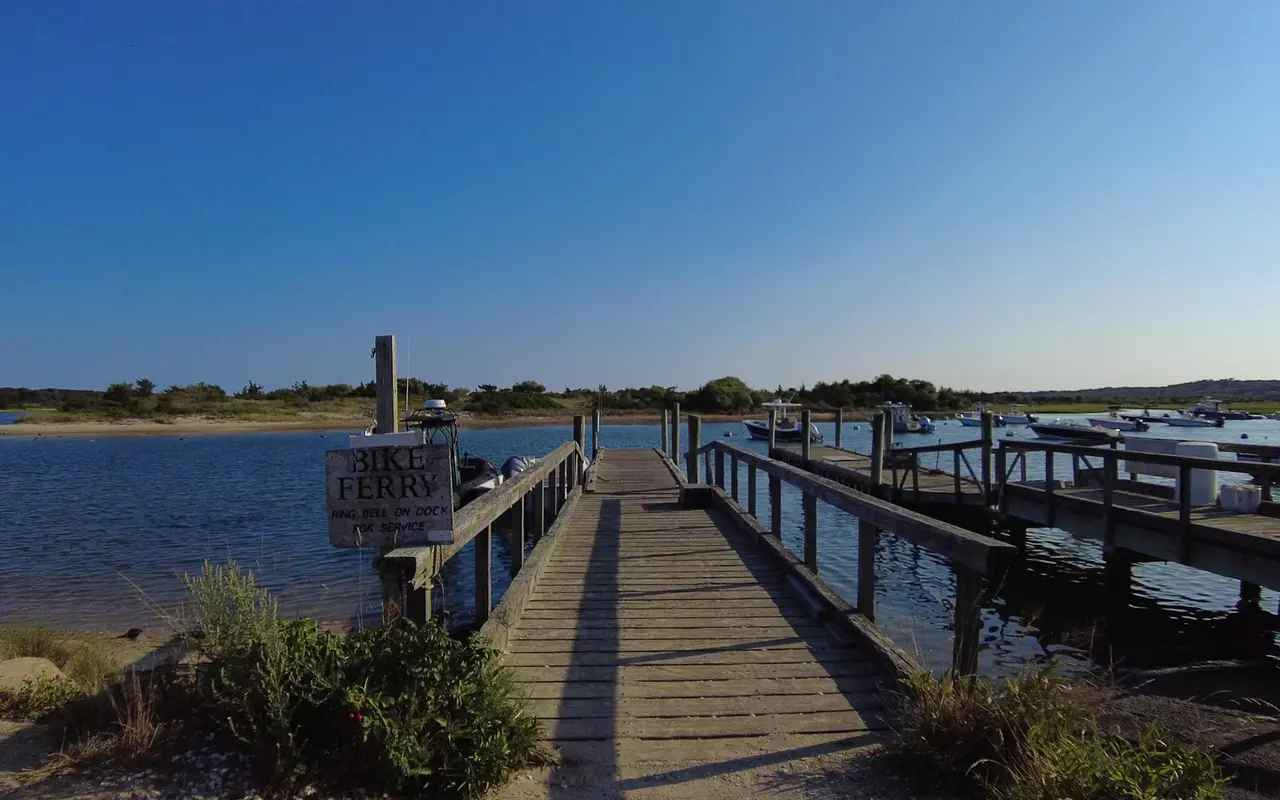 Wooden dock with a weathered Bike Ferry sign saying ring bell on dock for service, small boats moored in the channel and a sandy shore across the water