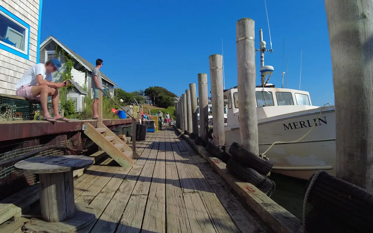 A white fishing boat named Merlin tied to tall pilings beside a long plank dock, shingled harbor shacks on the left and people sitting on the dock edge