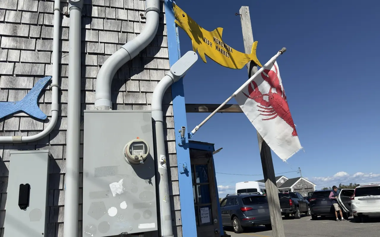 Weathered shingled wall of Menemsha Fish Market with a yellow shark sign and a lobster flag, cars in the parking lot beyond