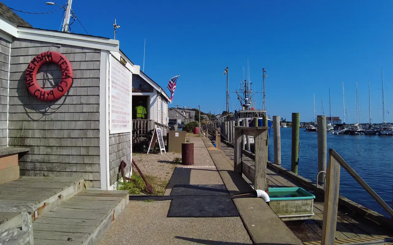 Red life ring reading Menemsha Texaco on a gray shingled building by the harbor, a flag, a fishing boat at the dock and sailboat masts in the harbor