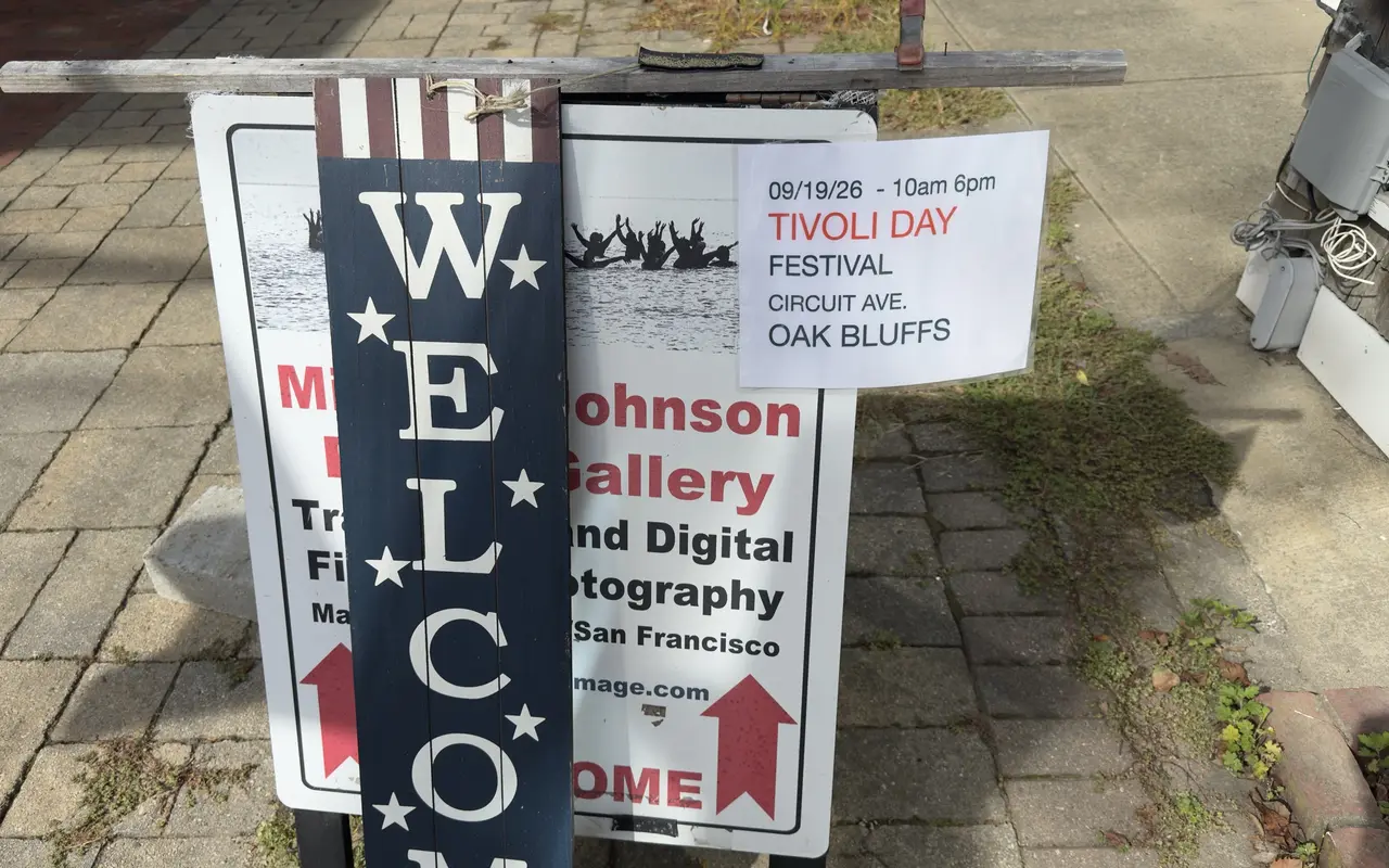 A sandwich board on a brick sidewalk in Vineyard Haven for the Michael Johnson Gallery, traditional and digital photography, with a wooden Welcome plank hung over it and a printed notice for the Tivoli Day festival on Circuit Avenue, Oak Bluffs, September 19, 2026, 10 to 6