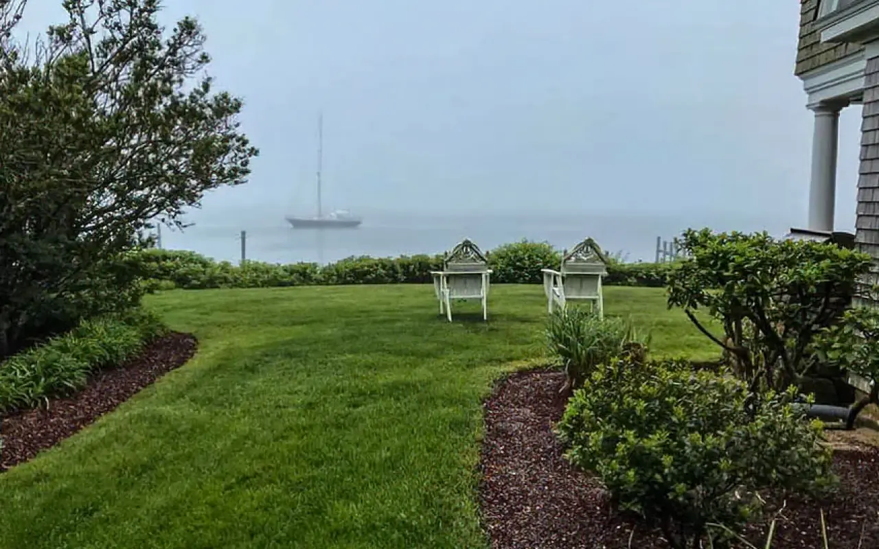Fog over a harbor lawn with two empty chairs and a moored sailboat