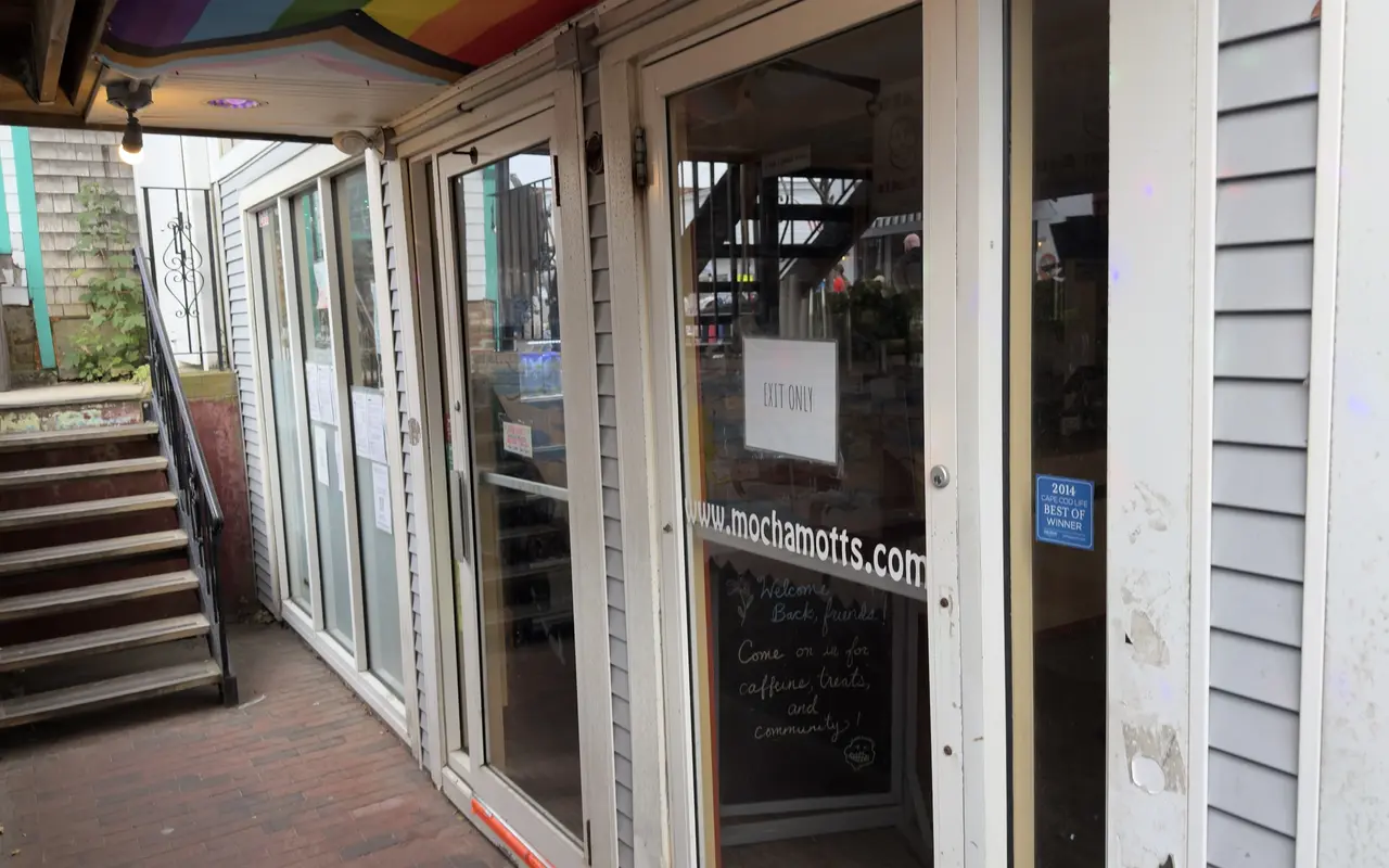 The glass door of Mocha Mott's in Oak Bluffs with an Exit Only sign, mochamotts.com lettering, a 2014 Cape Cod Life Best Of sticker, a chalkboard inside reading Welcome Back Friends, Come on in for caffeine, treats and community, and the stairs up to Circuit Avenue on the left
