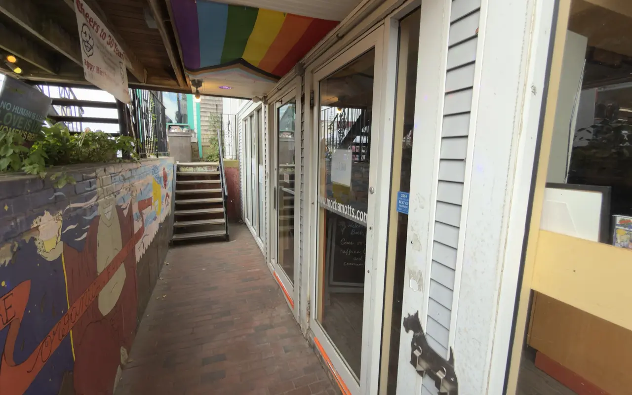 The basement walk to Mocha Mott's in Oak Bluffs: a painted mural along the low wall on the left, rainbow-striped awnings overhead, a Cheers to 30 Years banner, the glass door with mochamotts.com lettering and a chalkboard inside, stairs up to the street at the end