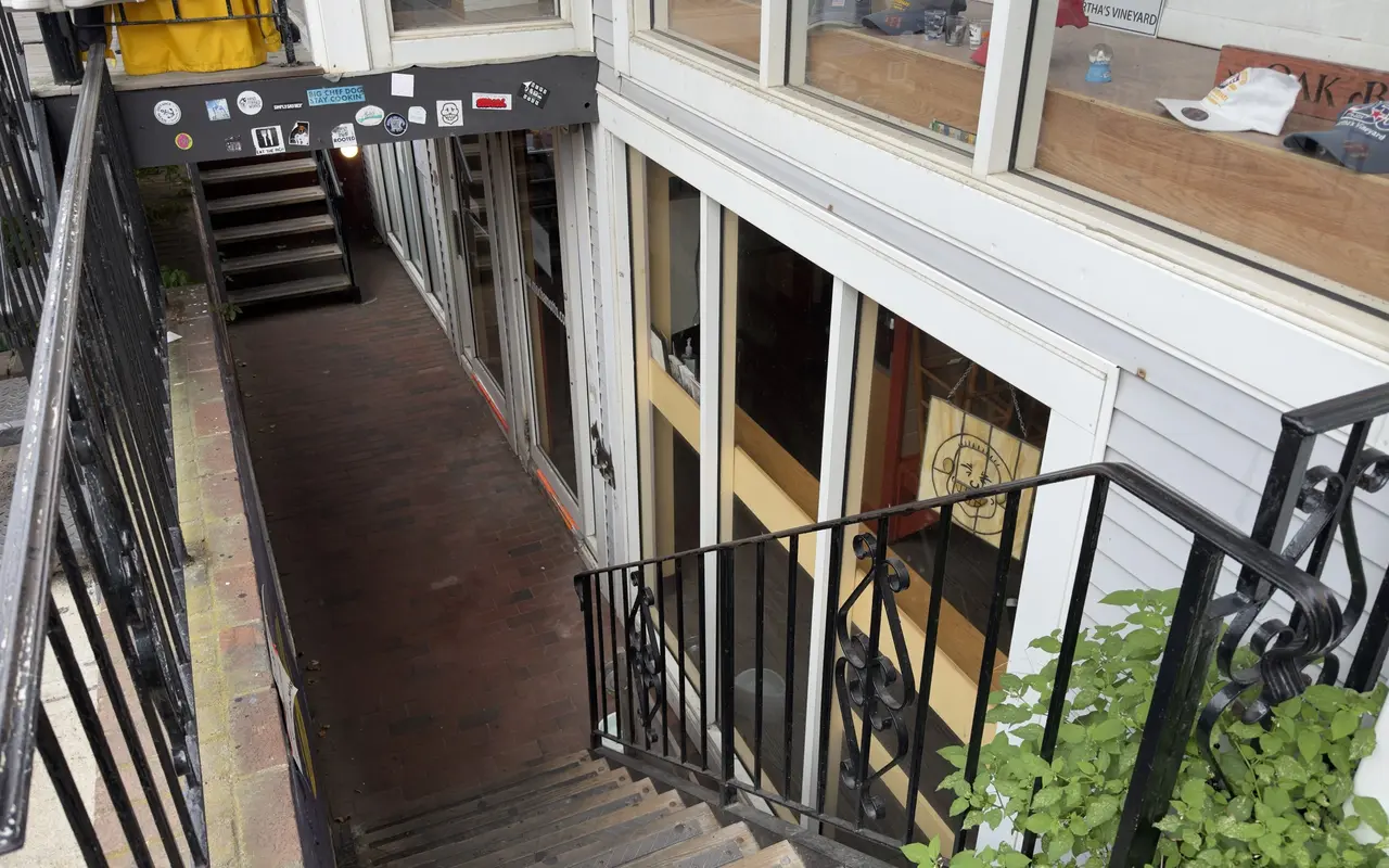 Looking down the iron stairs from Circuit Avenue to the basement walk of Mocha Mott's in Oak Bluffs: a brick passage between glass walls, a second stair at the far end, stickers along the beam, and Martha's Vineyard caps and an Oak Bluffs board in the shop window above