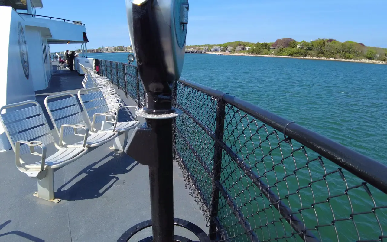 Rows of white metal seats along the railing of a ferry upper deck, a coin-operated binocular viewer in front and a wooded shoreline across blue water