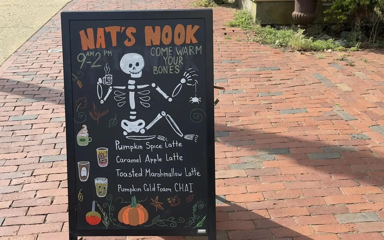 A chalkboard easel for Nat's Nook on a brick sidewalk in Vineyard Haven: hours 9 to 2, a skeleton holding a coffee, Come Warm Your Bones, and a fall list of pumpkin spice latte, caramel apple latte, toasted marshmallow latte and pumpkin cold foam chai, with pumpkins and leaves drawn around it