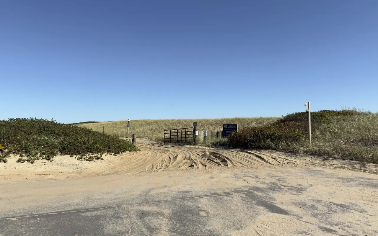 Sandy vehicle entrance to Norton Point Beach with deep tire tracks, a metal gate, a No Alcohol sign and a blue Welcome to Norton Point Beach sign listing four-wheel drive only, speed limit 15, deflate tires and permits required, dunes with beach grass behind