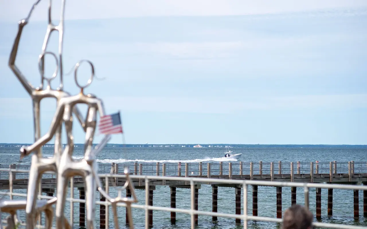 Stainless steel figure sculpture with a small American flag in front of a wooden pier and a passing motorboat