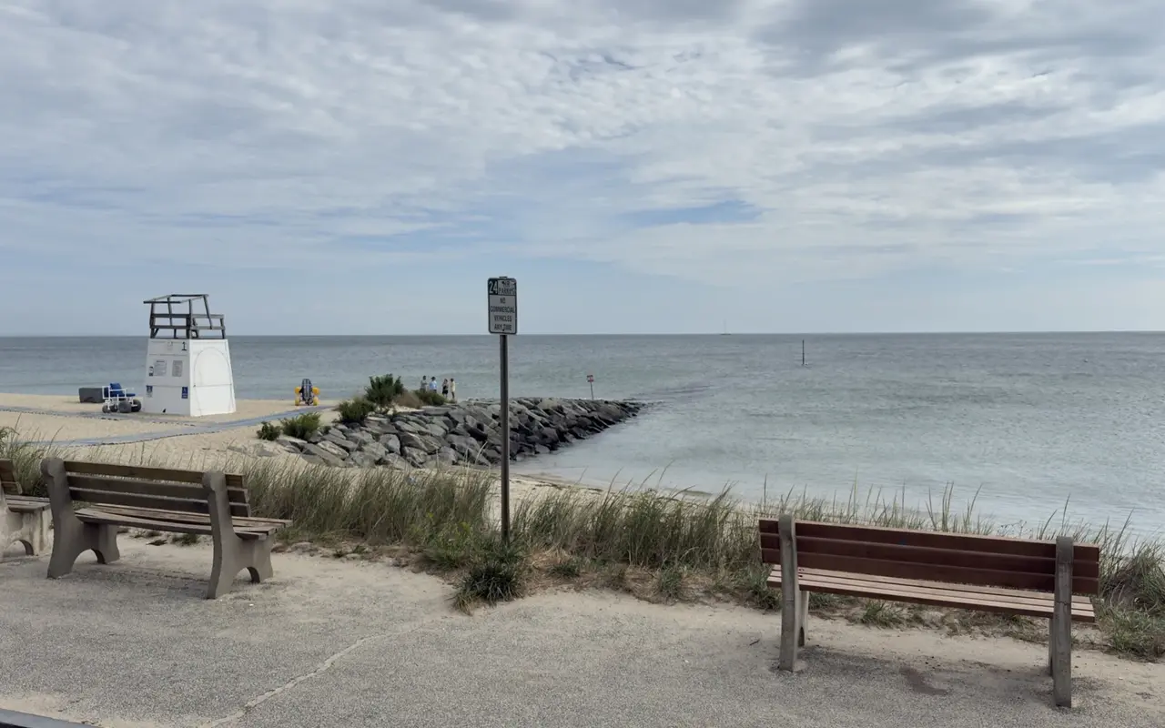 Benches on the walk above an Oak Bluffs beach, dune grass, a 24-hour parking sign, a lifeguard stand numbered 1 with a beach wheelchair and a yellow balloon-wheeled chair on the sand, a mat to the water and a rock jetty into flat, silvery water