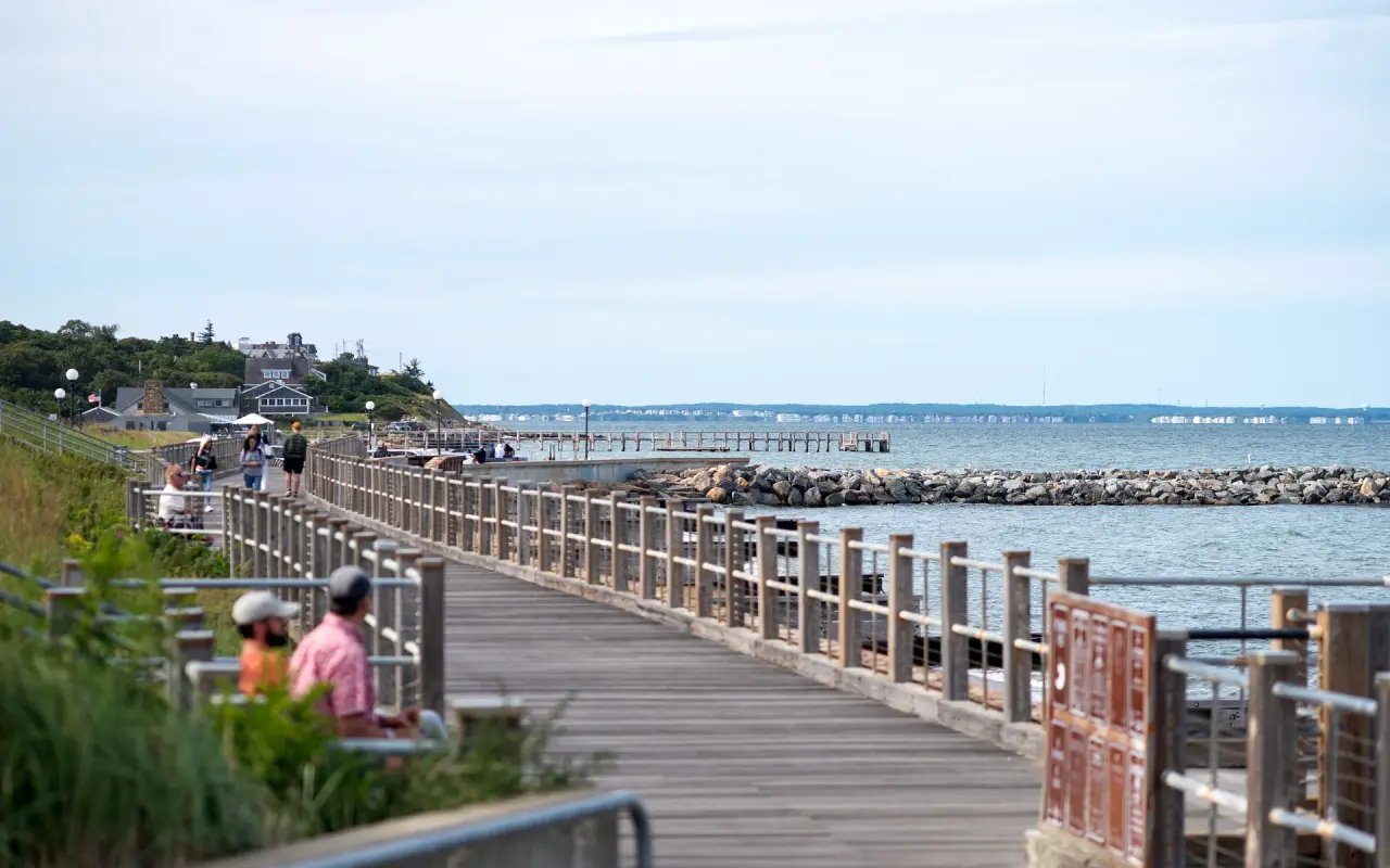 Wooden boardwalk along the Oak Bluffs shore with people on benches and a rock breakwater in the water