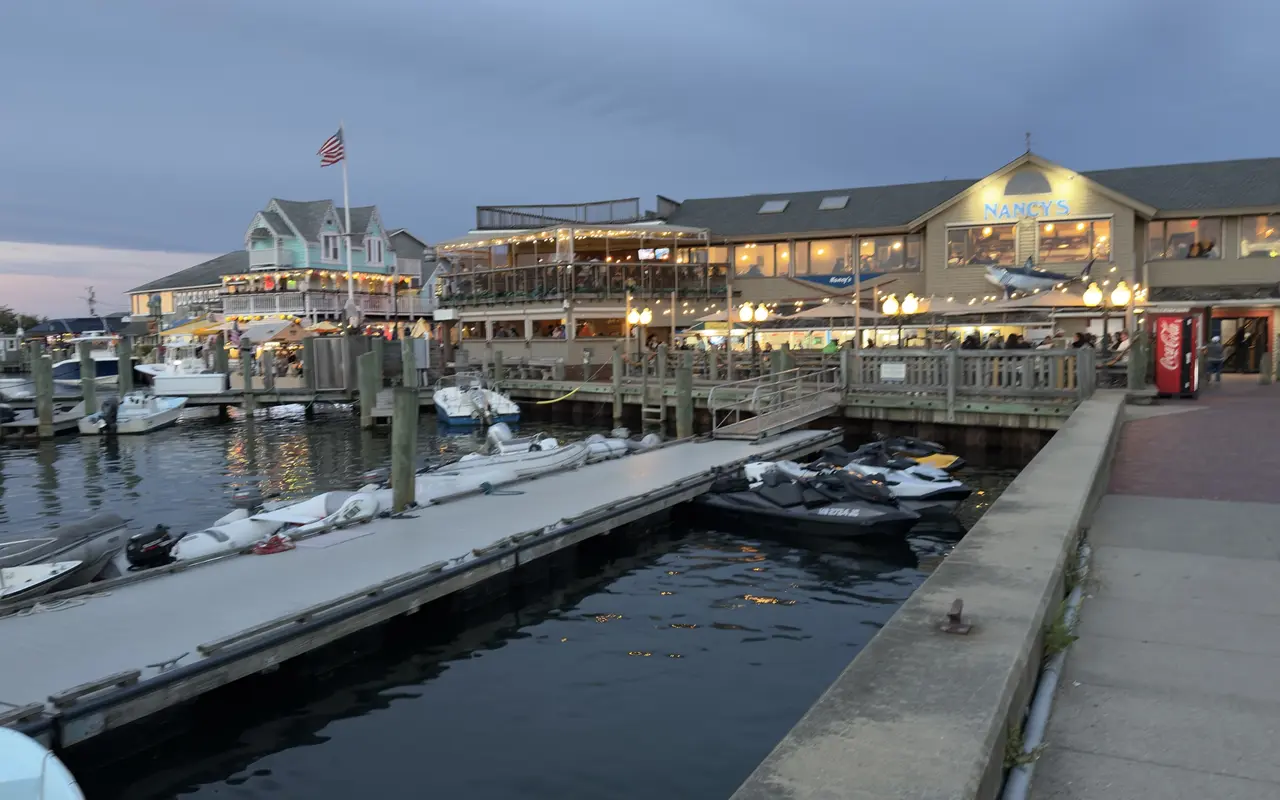 Oak Bluffs harbor at dusk: floating docks with jet skis and dinghies in the foreground, Nancy's restaurant lit up with its shark over the sign, the upstairs deck of the building next door strung with lights, a flag on a pole and a gingerbread house behind