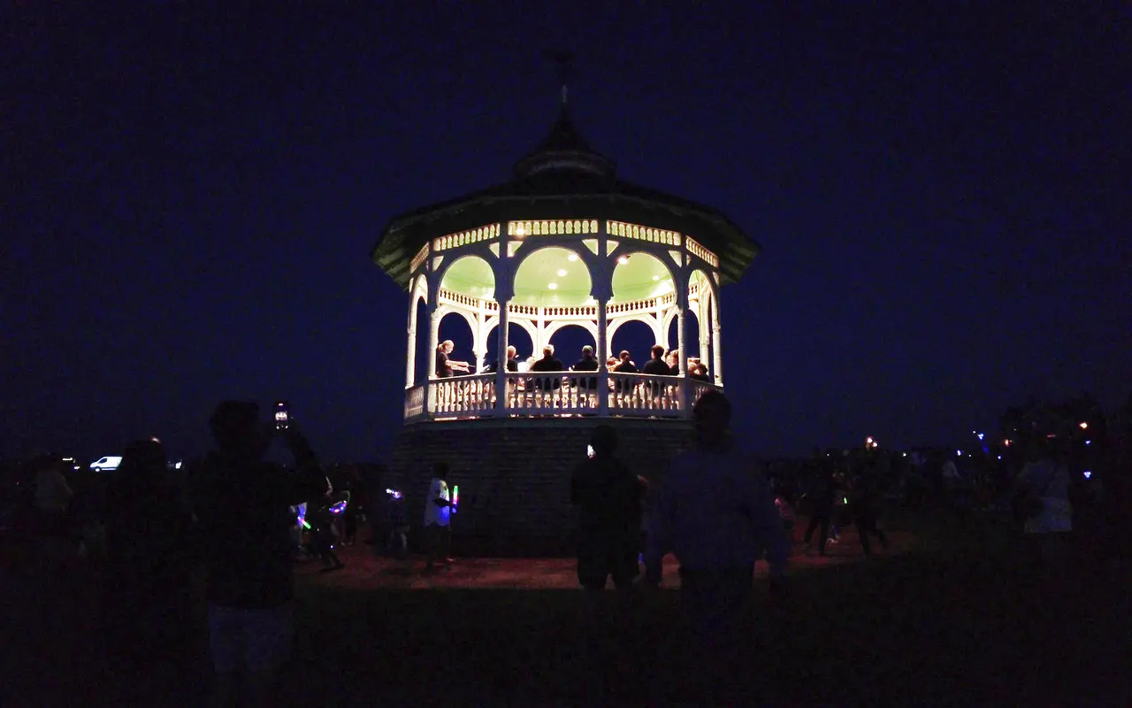The white Victorian bandstand in Ocean Park lit from inside at night, musicians on the platform and a crowd in silhouette on the lawn