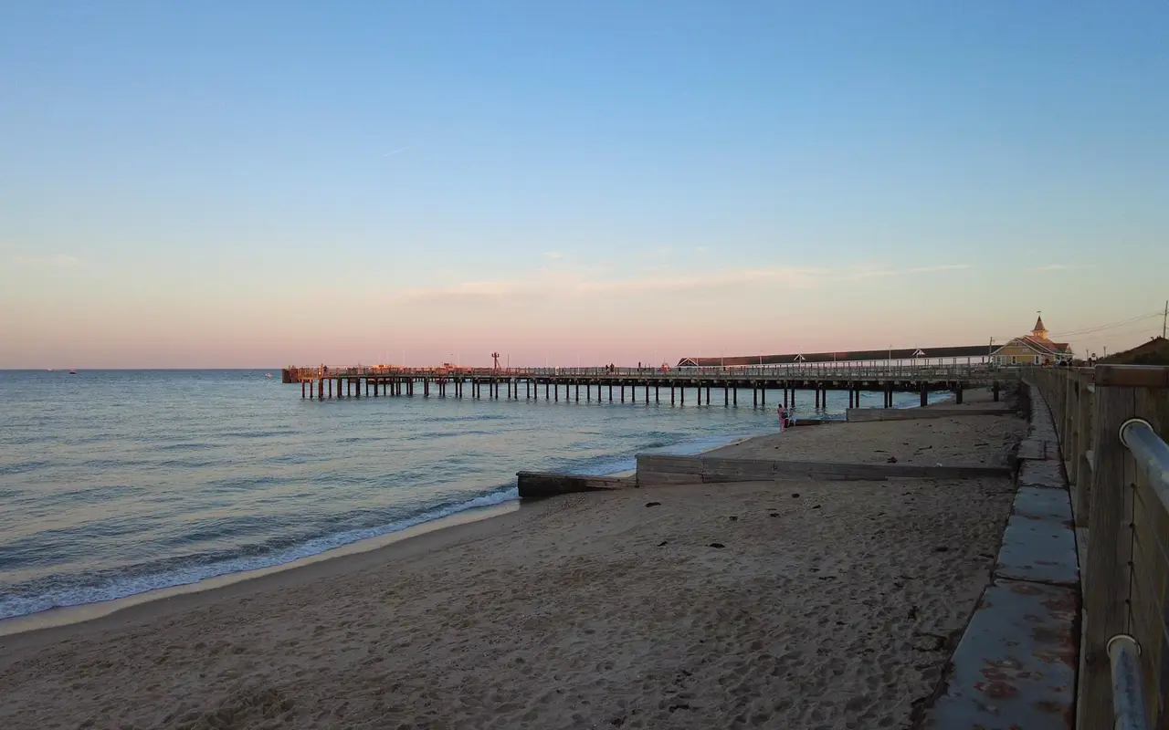 A long wooden pier reaching into calm water at dusk, a sandy beach below a seawall walkway and a building with a cupola at the shore end