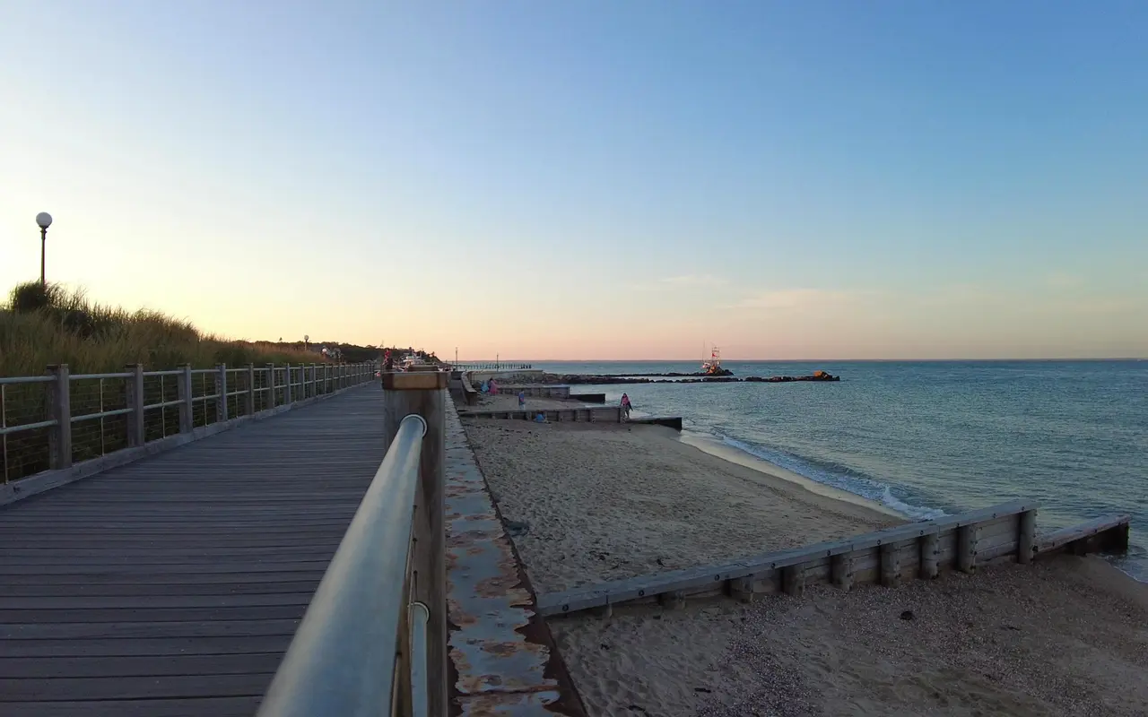 Wooden boardwalk with a metal railing above a sandy beach split by wooden groins, a stone jetty and calm sea under a pink dusk sky