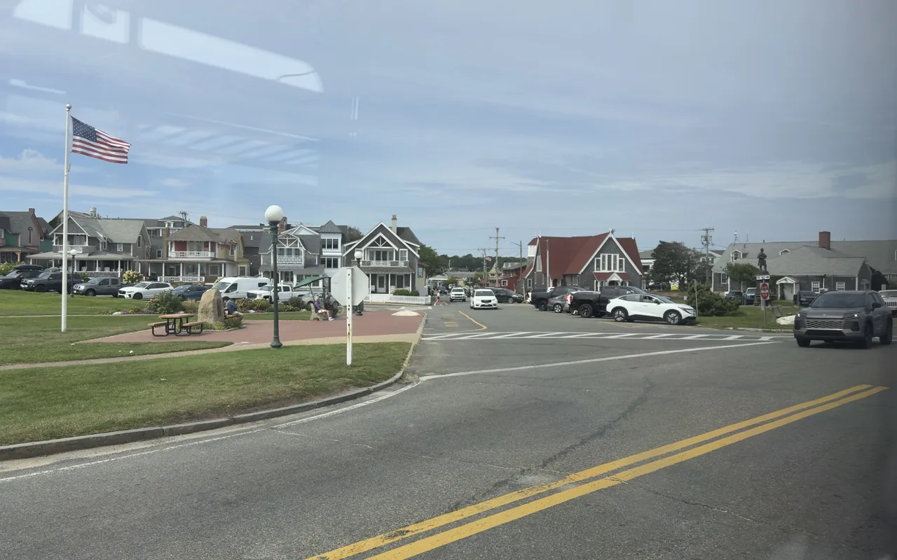 Ocean Park in Oak Bluffs seen through a bus window: the flagpole, benches, a stone marker and the shingled cottages along the park