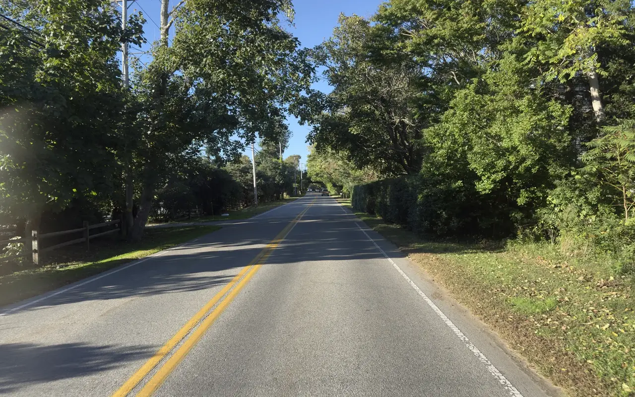 Old County Road in West Tisbury, a two-lane road with a double yellow line under a canopy of green trees, a split-rail fence on the left and a tall hedge on the right