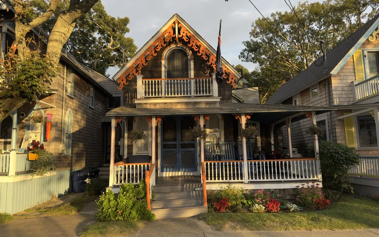 A shingled cottage in the Oak Bluffs Campground with orange scrollwork on the gable, an arched upstairs window, a white balcony and blue porch doors, in low, warm sunlight