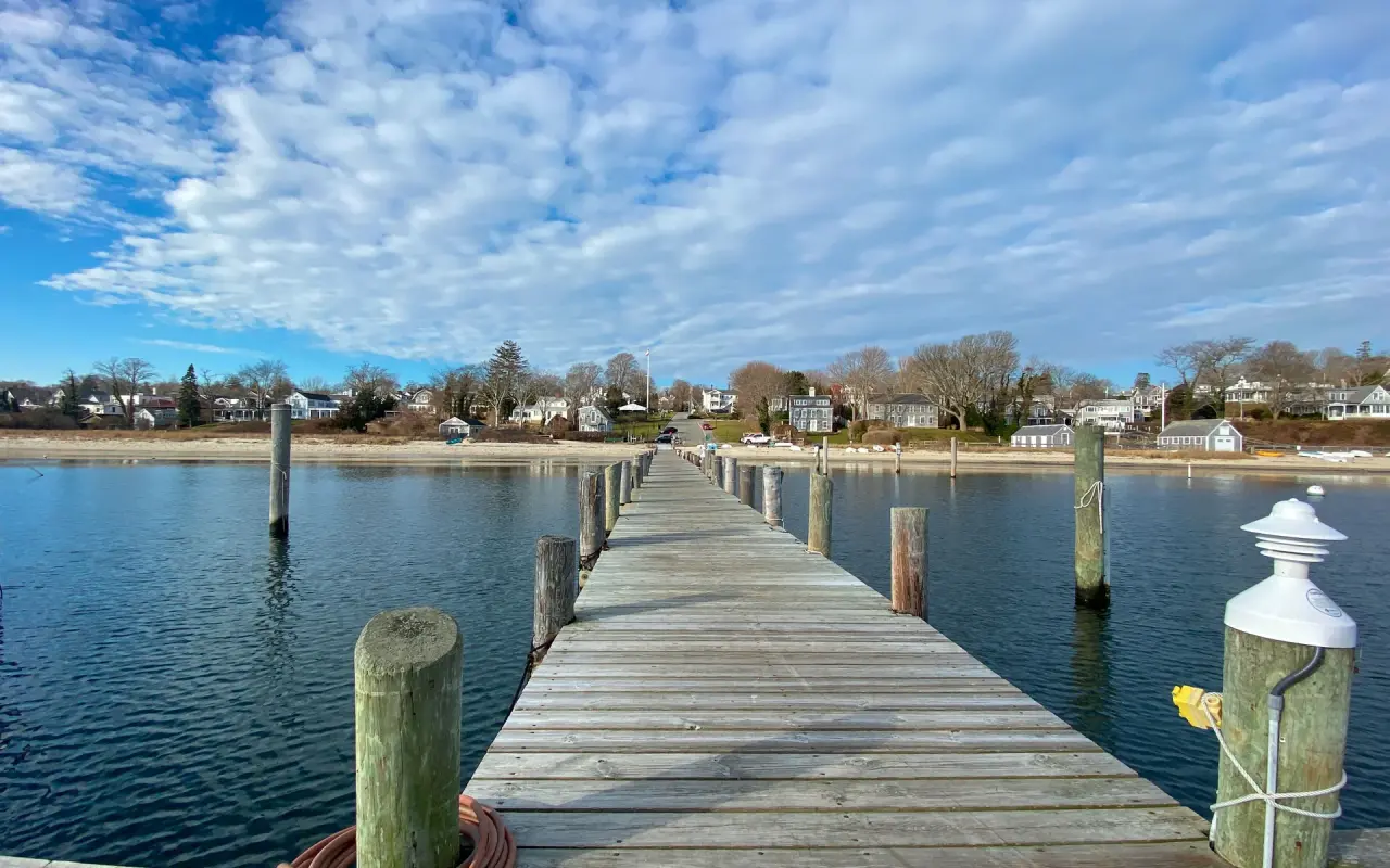 A wooden pier at Owen Park in Vineyard Haven under a blue sky with clouds, the harbor and houses beyond
