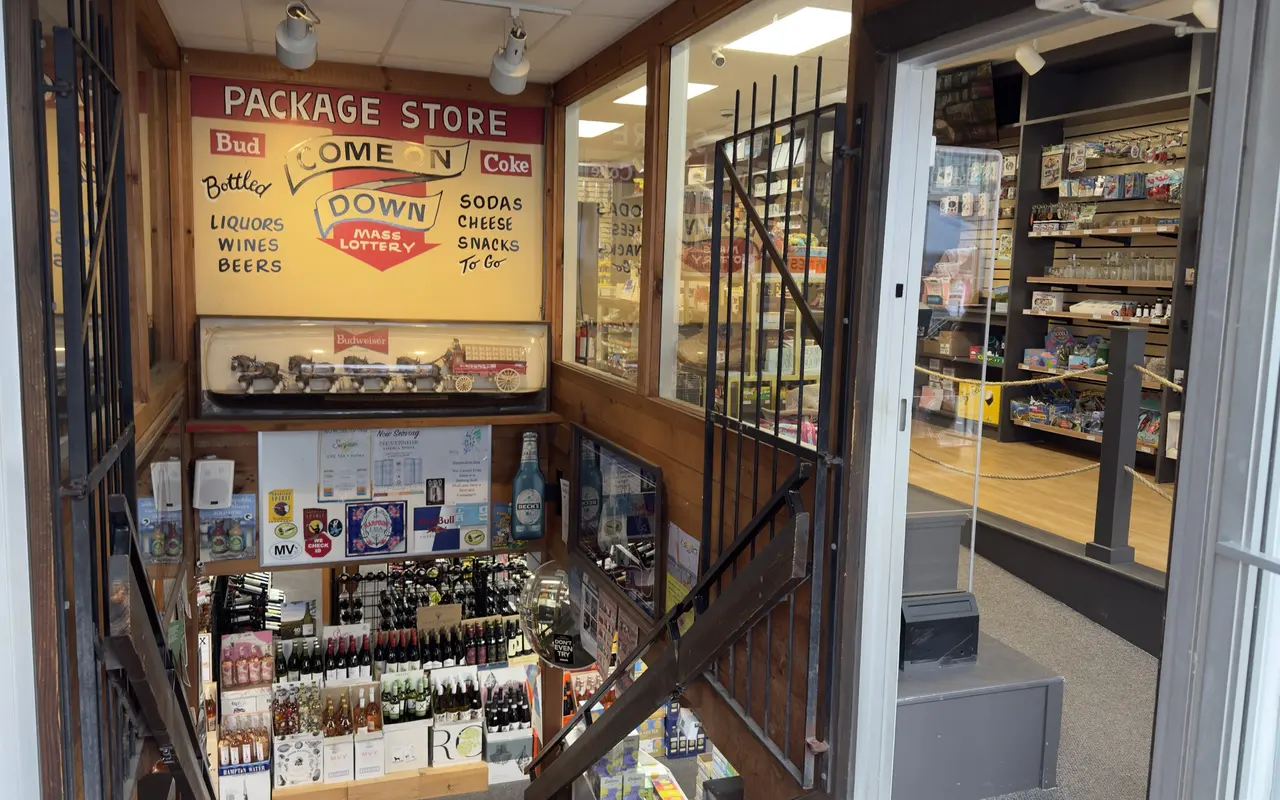 Looking down the stairs into a basement package store in Oak Bluffs: a hand-painted Package Store, Come On Down sign with Bud and Coke logos, bottled liquors, wines, beers, sodas, cheese and snacks to go, a Budweiser Clydesdale display, wine racks below, and a gift shop with mugs and souvenirs at street level beside it