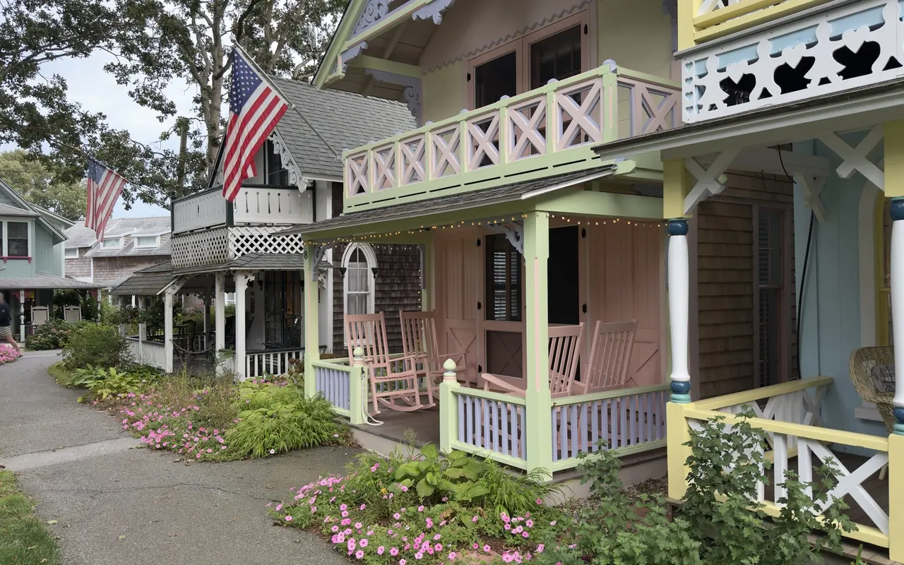 A row of gingerbread cottages in the Oak Bluffs Campground: a pink and pale green cottage with a balcony of X-pattern railings, pink rocking chairs and string lights on the porch, a yellow and blue cottage with heart cutouts beside it, American flags, pink flowers along the path