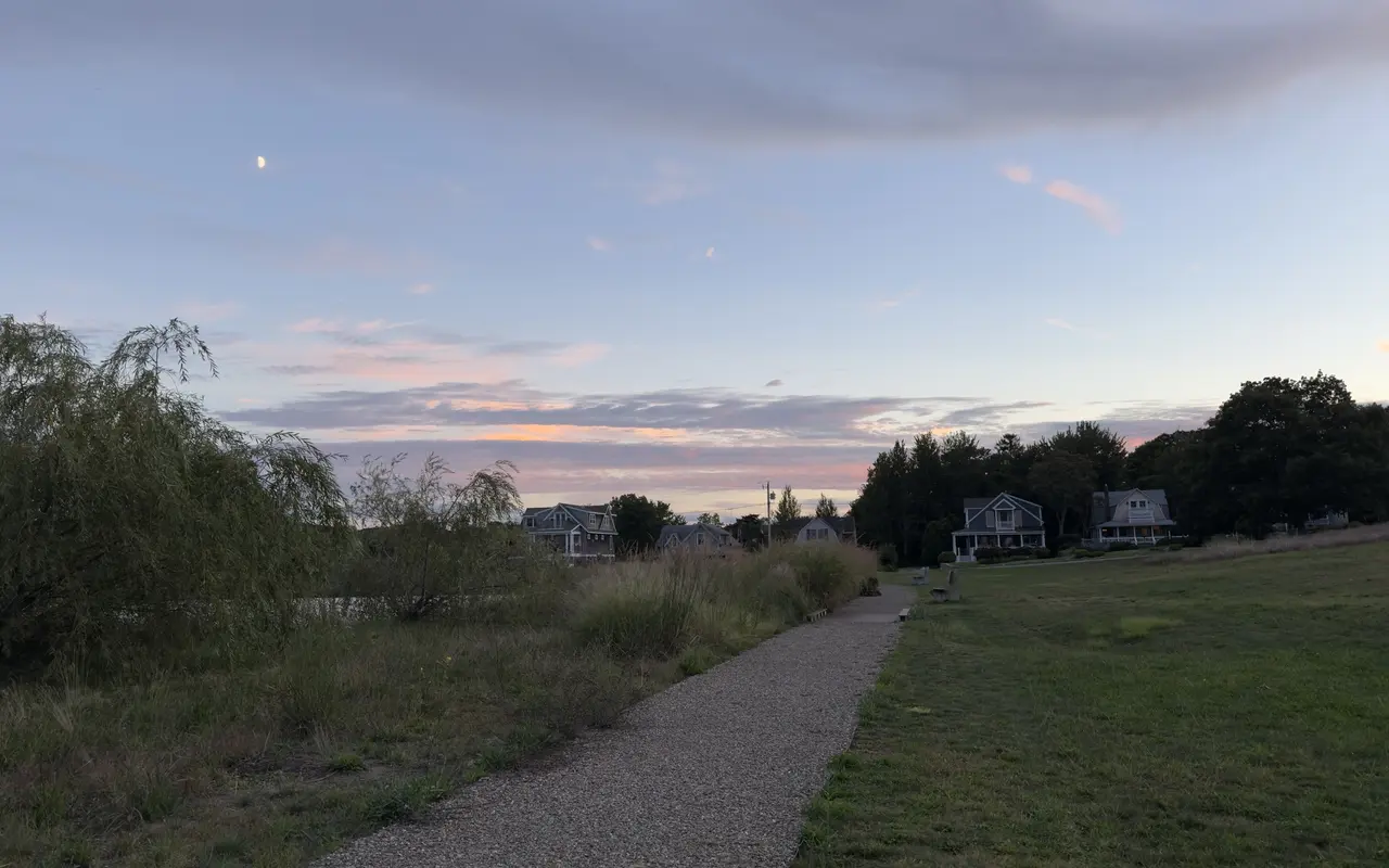 A gravel path beside a pond in Oak Bluffs at dusk, willows on the left, a lawn and shingled houses ahead, and a half moon in a pink-streaked sky