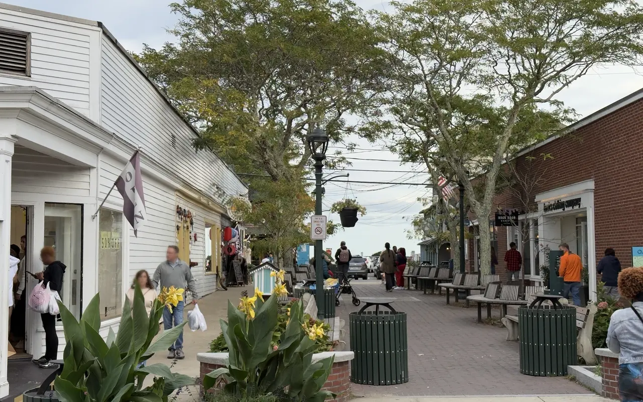 Post Office Square in Oak Bluffs on a September afternoon: brick paving, green slatted bins, benches under the trees, canna lilies in a planter, shoppers on the sidewalk by a 50% off sign, the Back Door Donuts sign on the brick building to the right and the Sound at the end of the square