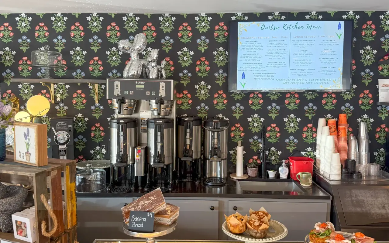 A café counter at Quitsa Kitchen in Vineyard Haven with coffee dispensers, pastries and a menu board on floral wallpaper