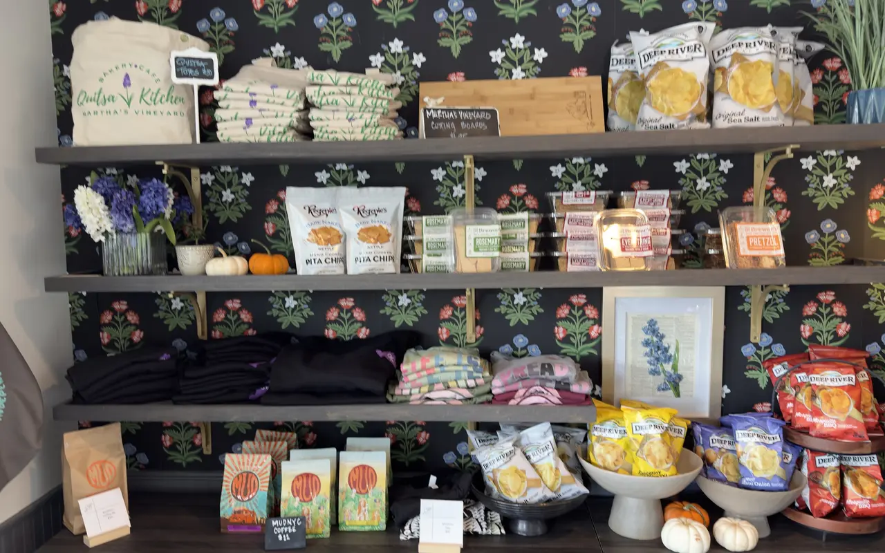 The retail wall at Quitsa Kitchen in Vineyard Haven: canvas totes with the bakery logo, island cutting boards, bags of Deep River chips, crackers, pita chips, folded hoodies and tees, a shelf of Mud coffee and small pumpkins, all against dark flowered wallpaper