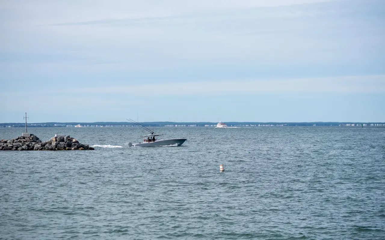 Center-console fishing boat with rods up passing a rock jetty, a ferry on the horizon