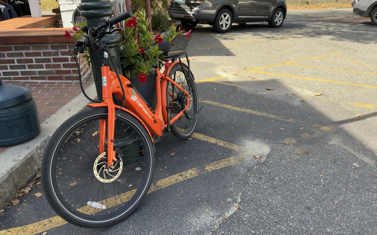 An orange step-through rental e-bike leaning against a lamppost and a planter of red flowers beside a parking lot